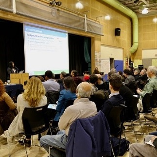 Many people sit in an auditorium. A DOT engineer presents information on a large screen