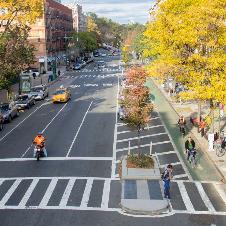 A wide, tree-lined one-way street in NYC with three vehicle lanes, a a wide concrete median, a green bike lane, and a sidewalk.