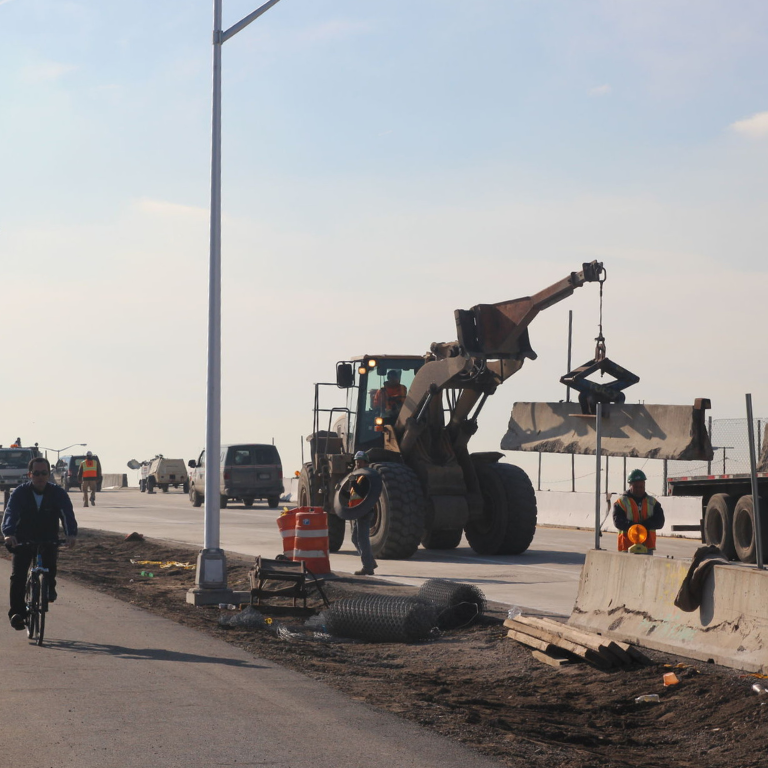 Construction vehicles move concrete jersey barriers on a wide roadway next to a bicycle path