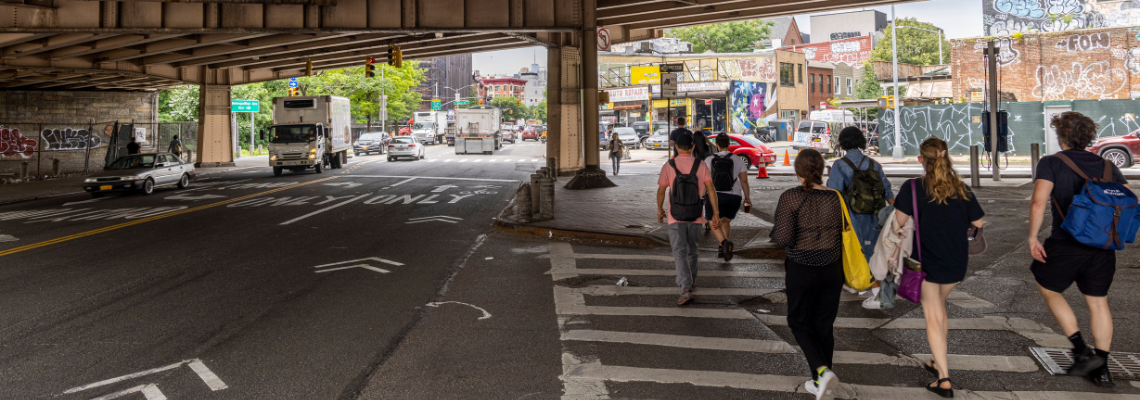 A group of people walk across a wide street under an elevated roadway in Brooklyn